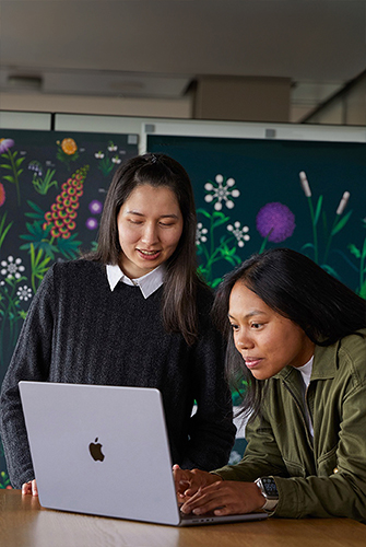 Two Apple Marketing team members working together on a MacBook in front of posters designed by their team.