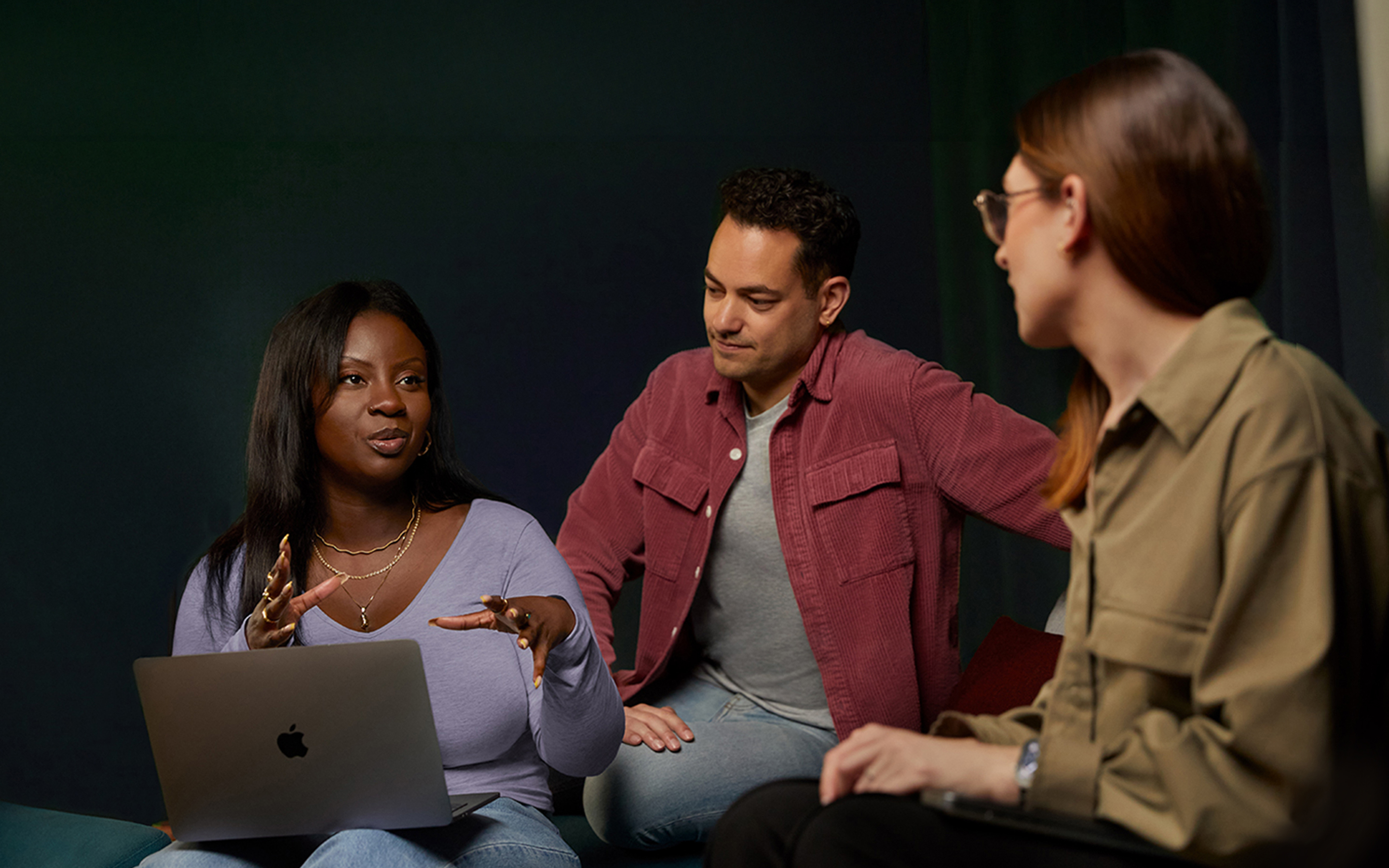 Three Apple Software and Services team members talking together on a couch, one with a MacBook.