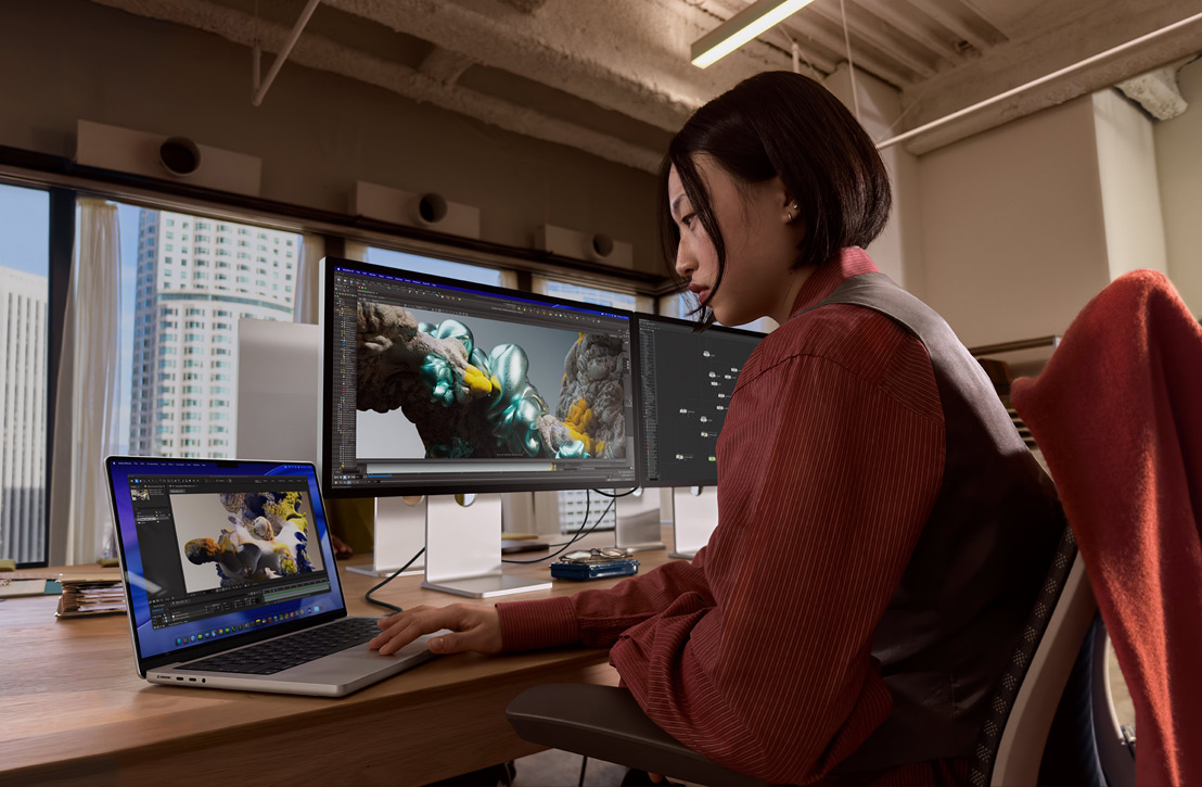 Eine Person arbeitet an einem Schreibtisch mit einem MacBook Pro, das mit einem externen Display verbunden ist, beide Displays zeigen ein 3D Rendering, im Hintergrund sind durch ein Fenster Gebäude zu sehen