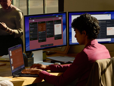Een team zit op kantoor rondom een tafel. Een vrouw zit achter haar bureau op een MacBook en displays te werken. Haar collega’s zitten erbij en zijn in gesprek.