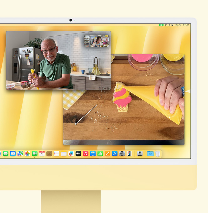 Ein iMac in Gelb. Eine Demonstration der Schreibtischansicht. Auf dem Bildschirm wird eine Bäckerin in einem Videoanruf mit einer anderen Person gezeigt. Auf einem Bildschirm werden die Bäckerin und das Gesicht des Mannes gezeigt. Auf dem anderen Bildschirm werden die Hände der Bäckerin beim Glasieren eines Kekses gezeigt