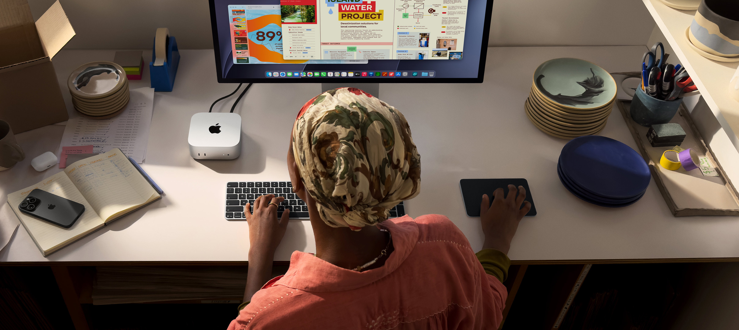 Henkilö kirjoittaa työpöydän ääressä Mac minillä, Studio Display liitettynä, Magic Keyboard, Magic Trackpad ja erilaisia esineitä