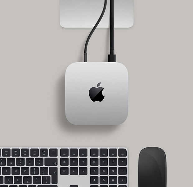 Mac mini, top, silver aluminum, rounded corners, glossy black Apple logo centered, Studio Display stand partially visible, Magic Keyboard with Touch ID, Magic Trackpad