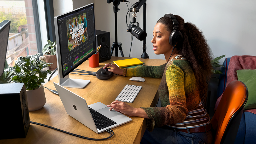 امرأة في منزلها تعمل خلف مكتبها على MacBook Pro متصل بشاشة خارجية وتستخدم سماعات رأس وتتحدث في الميكروفون