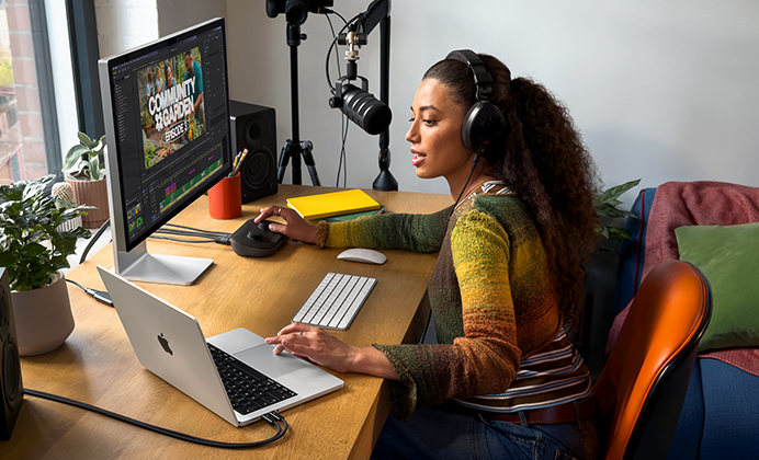 A woman in her home working at her desk with MacBook Pro connected to an external display, wearing headphones and speaking into a microphone