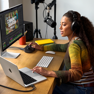 Evindeki çalışma masasında harici bir ekrana bağlı MacBook Pro ile çalışan bir kadın, kulaklık takıyor ve bir mikrofona konuşuyor