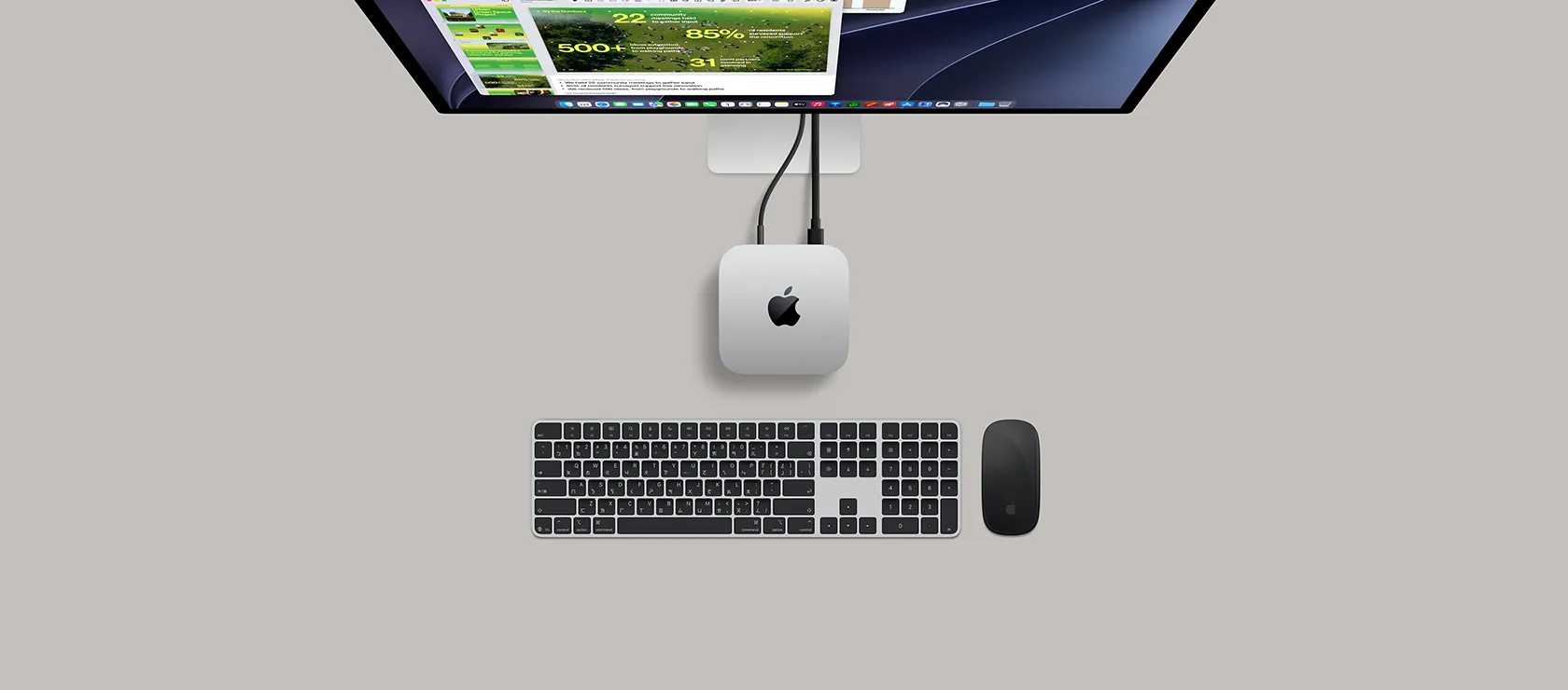 Mac mini 的俯視圖，展示銀色鋁金屬機身、圓角設計、機身中央的黑色亮面 Apple 標誌，透過連接線連接，頂部可見部分 Studio Display 機身，螢幕顯示行事曆 app、Keynote 簡報，以及打開的條形表格，Mac mini 下方可見含數字的巧控鍵盤與巧控滑鼠
