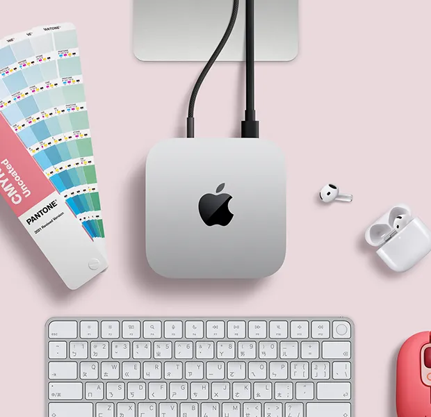 Mac mini 的俯視圖，展示銀色鋁金屬機身、圓角設計、機身中央的黑色亮面 Apple 標誌，透過連接線連接，Studio Display 支架部分可見，旁邊放著 AirPods 4、Pantone 色票，以及紅色的 Logitech 鍵盤與滑鼠