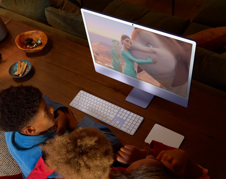 Tre unge sidder ved et bord med en iMac, Magic Keyboard og Magic Trackpad og ser en film