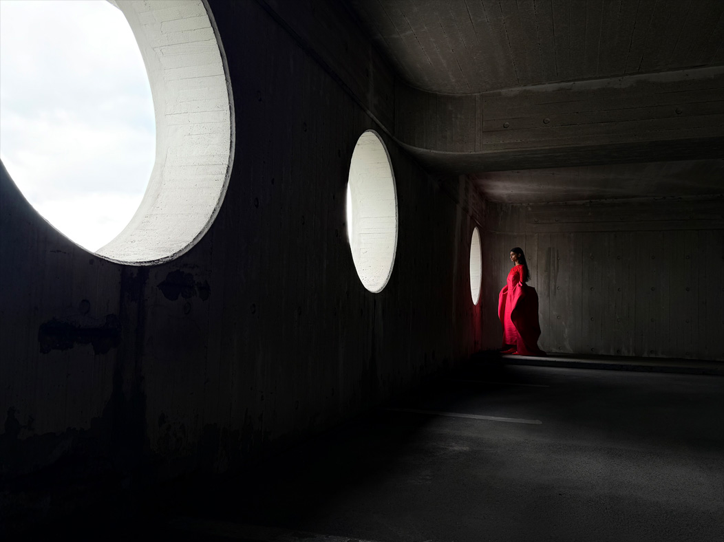 Uma mulher com um longo vestido vermelho a ser fotografada numa sala vazia. A app Fotografia muda de distância focal para ilustrar as capacidades do sistema de câmaras Pro, a mostrar a opção de 28 mm
