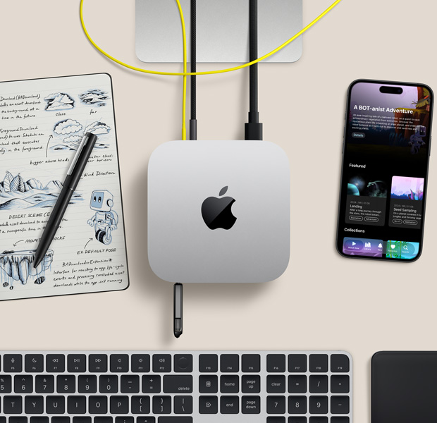 Mac mini, top, silver aluminum, rounded corners, glossy black Apple logo centered, connected by cord, USB stick inserted in USB-C port in front, Studio Display stand partially visible, iPhone, Magic Trackpad, Magic Keyboard with Touch ID, stylus and e-ink sketch pad