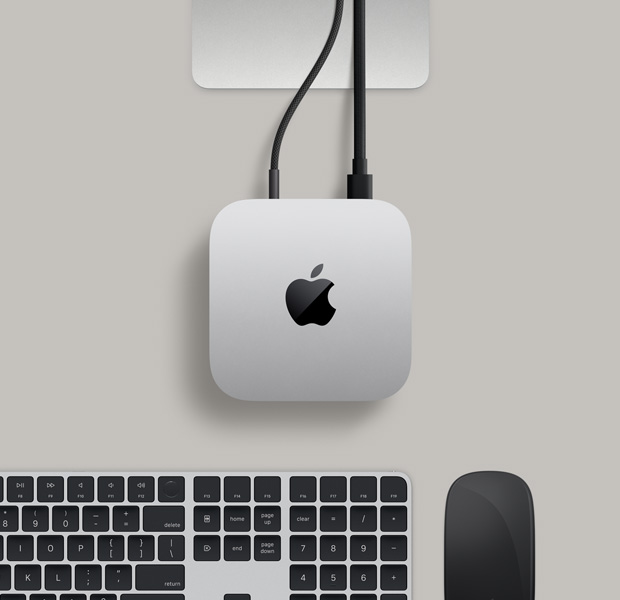 Mac mini, top, silver aluminum, rounded corners, glossy black Apple logo centered, Studio Display stand partially visible, Magic Keyboard with Touch ID, Magic Trackpad