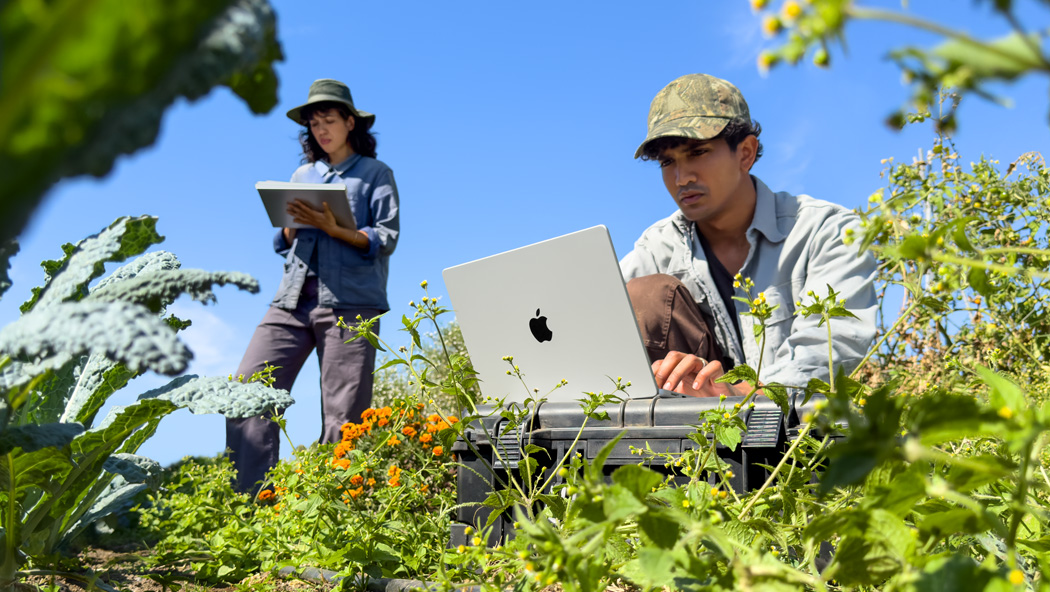 Zwei Personen arbeiten im Freien in einem Garten, eine nutzt ein MacBook Pro in Silber, das nicht am Strom angeschlossen ist, und die andere hält ein Notizbuch