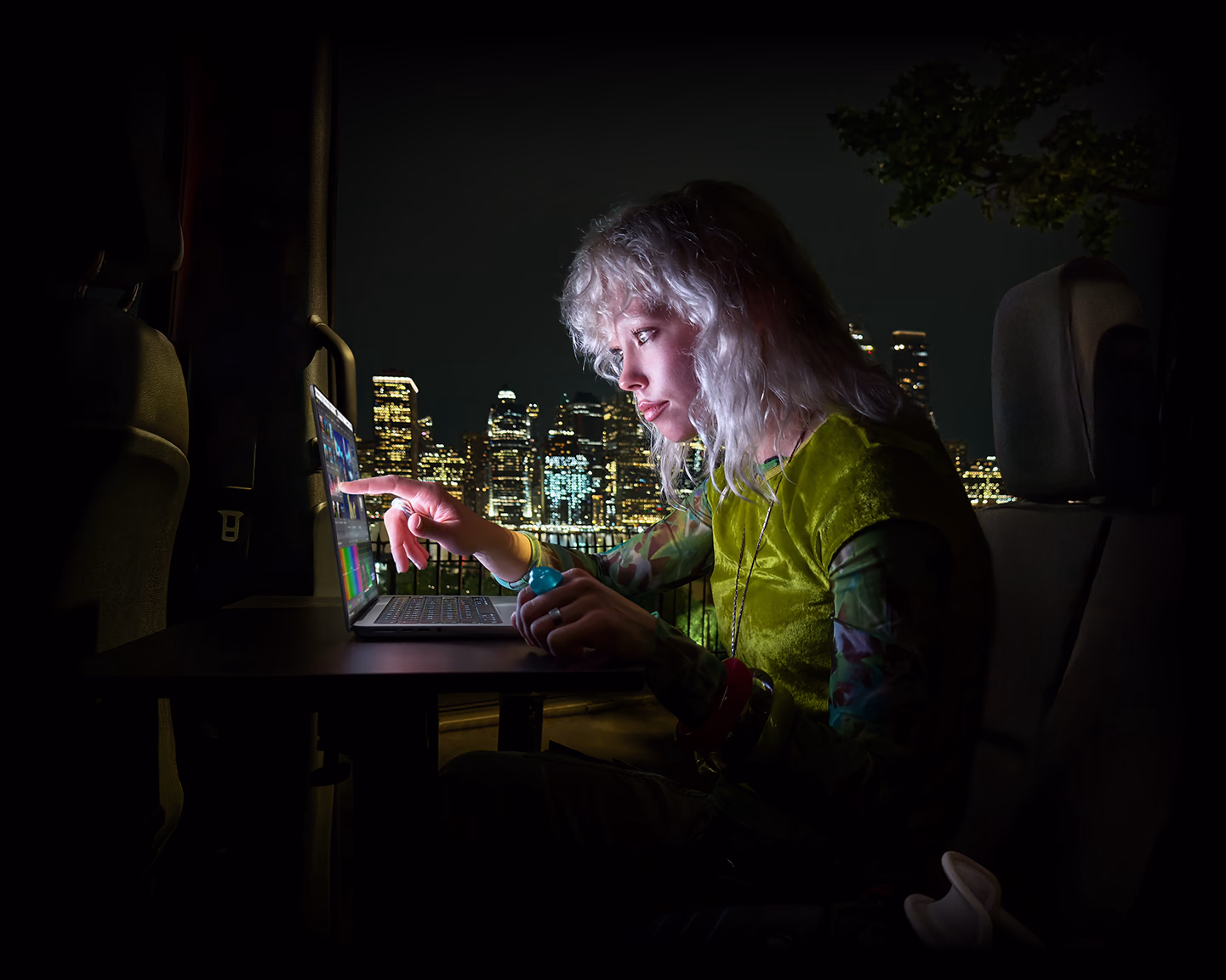 Eine Person sitzt auf dem Rücksitz eines Vans, durch die geöffnete Beifahrertür ist die Skyline einer Stadt bei Nacht zu sehen, ein MacBook Pro mit PROLED XDR Display auf einem Schreibtisch, die Person wird vom Bildschirm beleuchtet, während sie das Touch Feature nutzt, um ein Video in DaVinci Resolve Studio zu bearbeiten