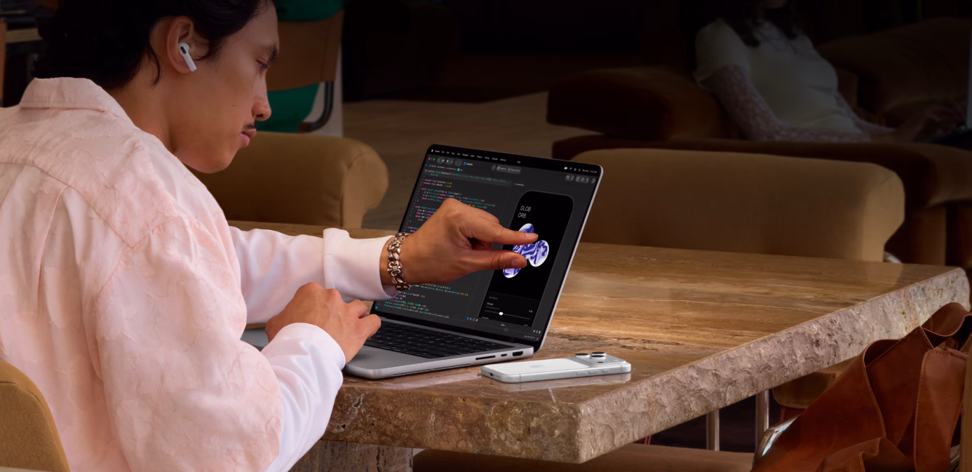 Eine Person sitzt an einem Tisch in einem öffentlichen Raum und passt mit einer Handgeste ein Element in Xcode an. Sie nutzt dabei das Touch Feature eines MacBook Pro in Silber mit PROLED XDR Display, neben dem Laptop befindet sich ein iPhone auf dem Tisch und die Person trägt AirPods Pro